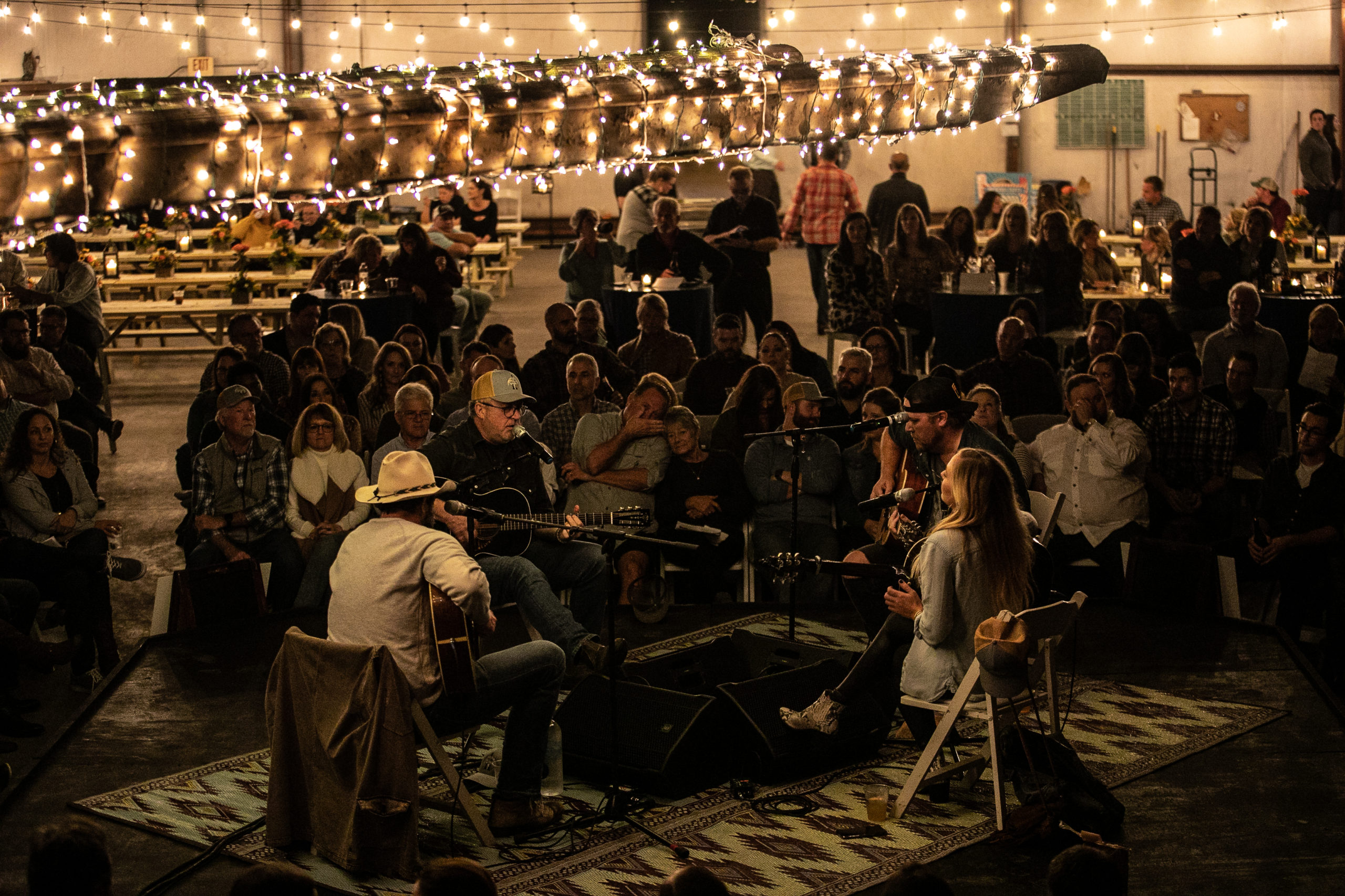 An intimate live Songwriters' Round with an engaged audience under the warm glow of string lights.